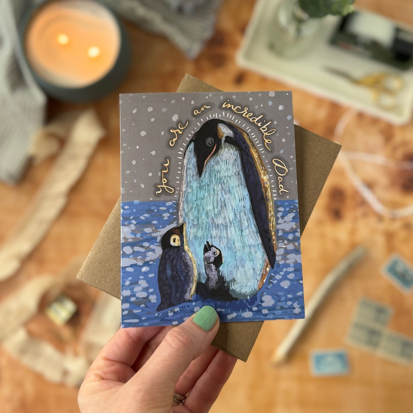 Hand holding a card with penguins and text on a wooden surface with a candle and plant.