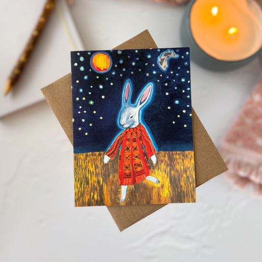 A greeting card featuring a whimsical illustration of a white rabbit in a quilted red coat under a starry night sky, styled on a kraft envelope with a pink blanket, candle, and notebook around it.