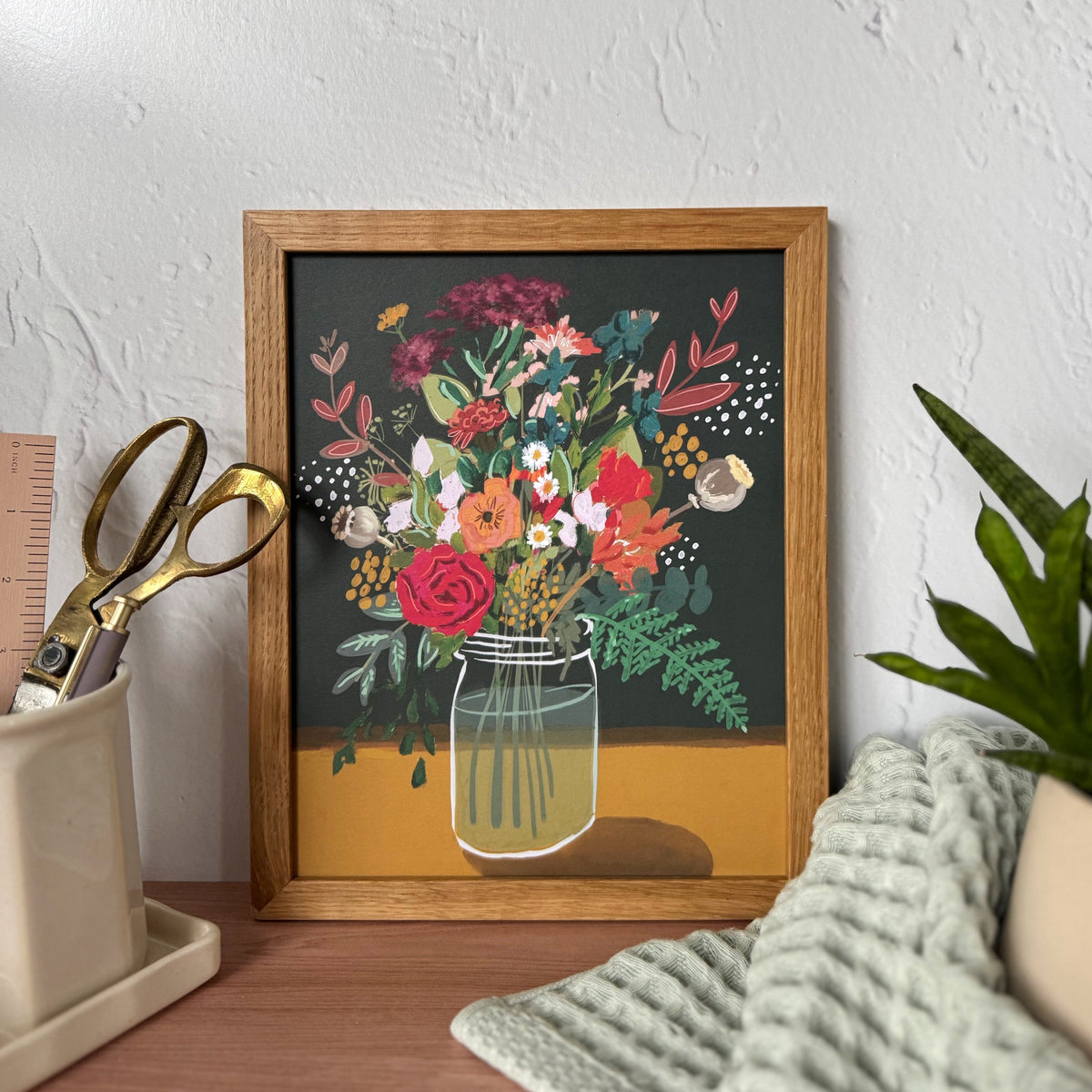 A framed floral art print featuring a colorful bouquet in a glass jar against a dark background, styled on a wooden desk with a plant, textured towel, and scissors in a cream holder against a textured white wall.