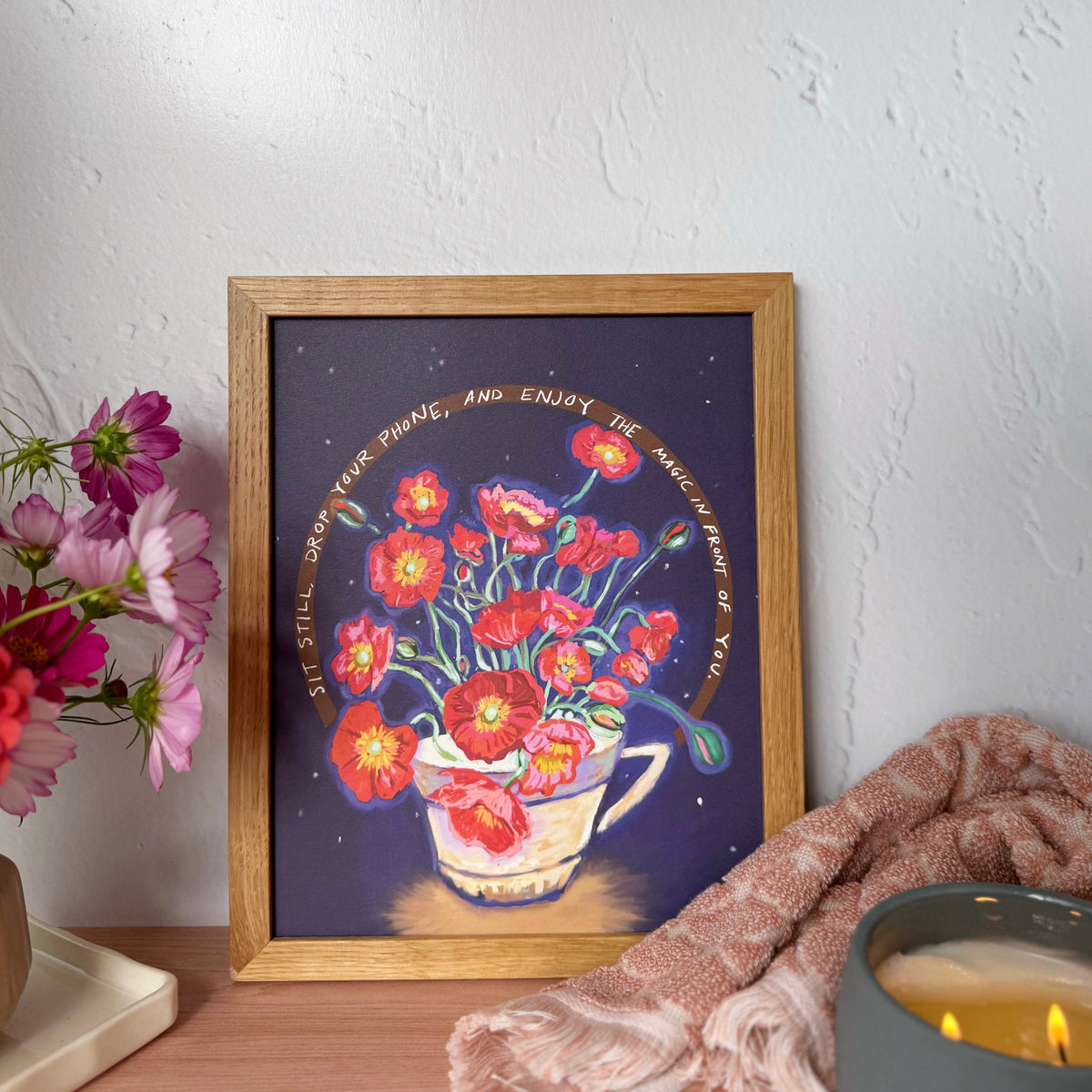 A framed art print featuring a whimsical illustration of red poppies in a teacup against a dark purple background, with words that say, "Sit still, drop your phone, and enjoy the magic in front of you." Styled on a wooden table with a pink towel, a lit candle, and flowers against a textured white wall.