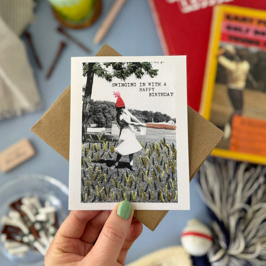 Hand holding a birthday card with a black and white photo of a woman golfing in a field, surrounded by various items on a table.