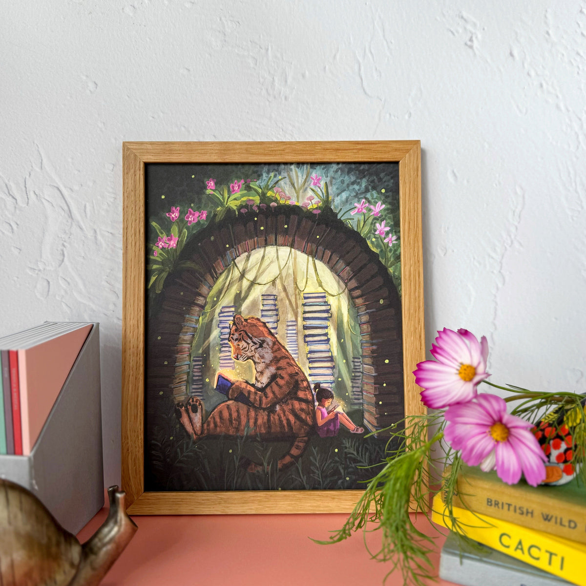 A framed art print featuring a tiger and little girl reading, sitting among arched books and flowers, styled on a pink surface against a white textured wall with decorative books and flowers.