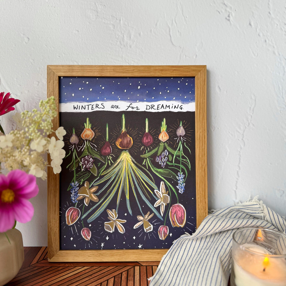 A framed art print featuring whimsical botanical illustrations of flowers and bulbs under a dark blue snowy sky with the text 'WINTERS are FOR DREAMING'. The print is framed in light wood and styled on a wooden surface next to a candle, a striped cloth, and flowers against a textured white wall.