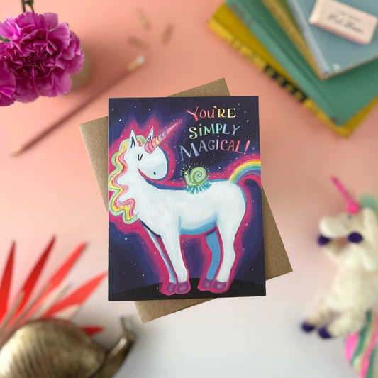 A square greeting card featuring a colorful unicorn and snail illustration with the text 'You're Simply Magical!' on a dark starry background, styled on a kraft envelope with flowers, books, and a plush toy around it.