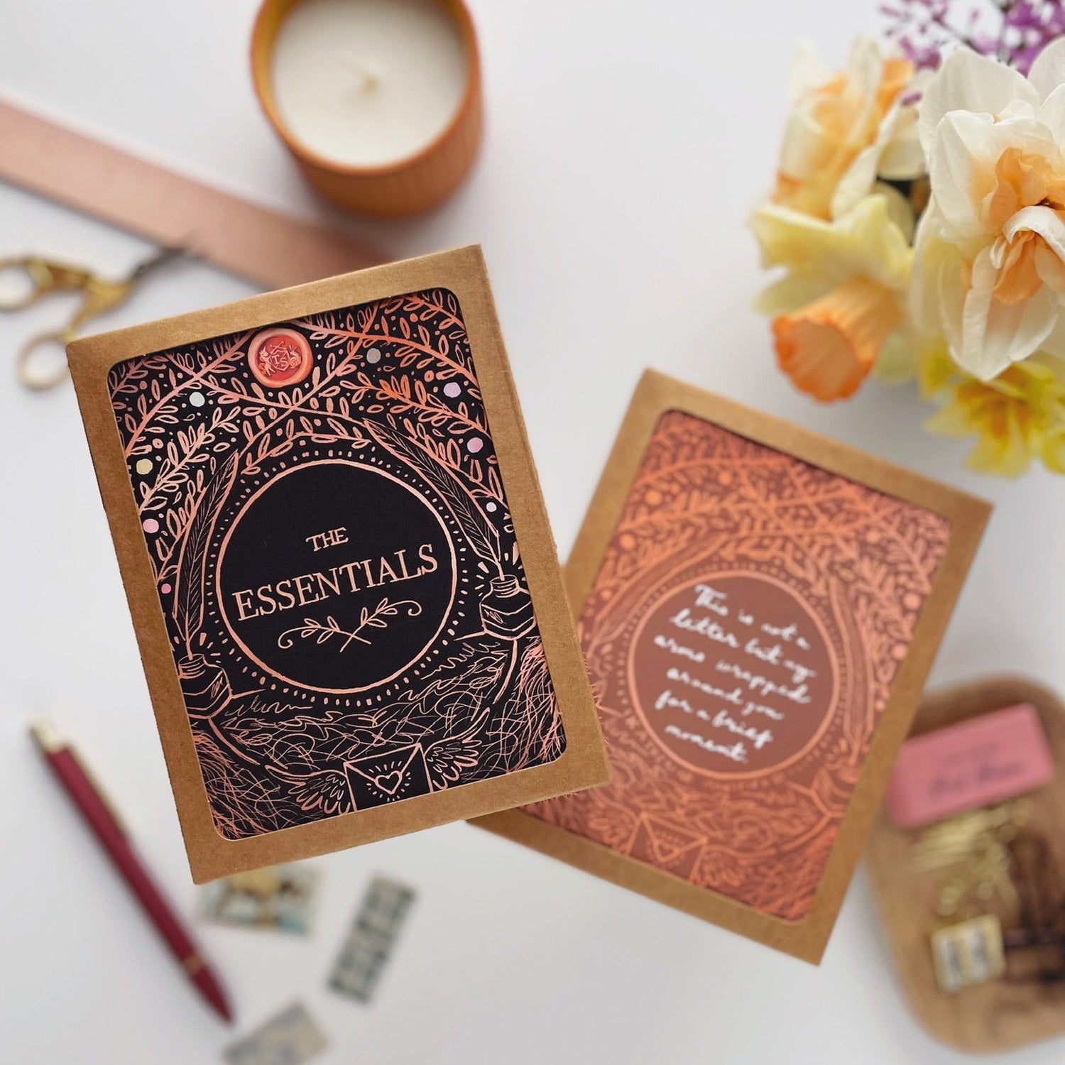 Two decorative boxes with intricate designs and text, placed on a light surface with a candle and flowers in the background.