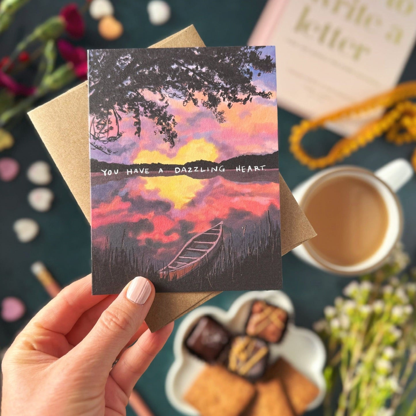 Hand holding a card with a scenic design and text that says "You have a dazzling heart," surrounded by cookies, coffee, and flowers.