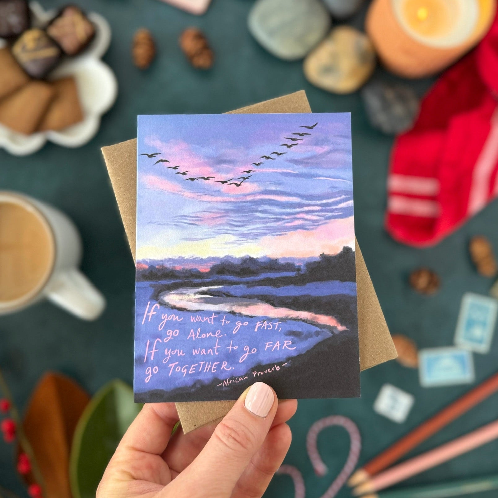 Hand holding a greeting card with a scenic design and text, surrounded by decorative items on a table.