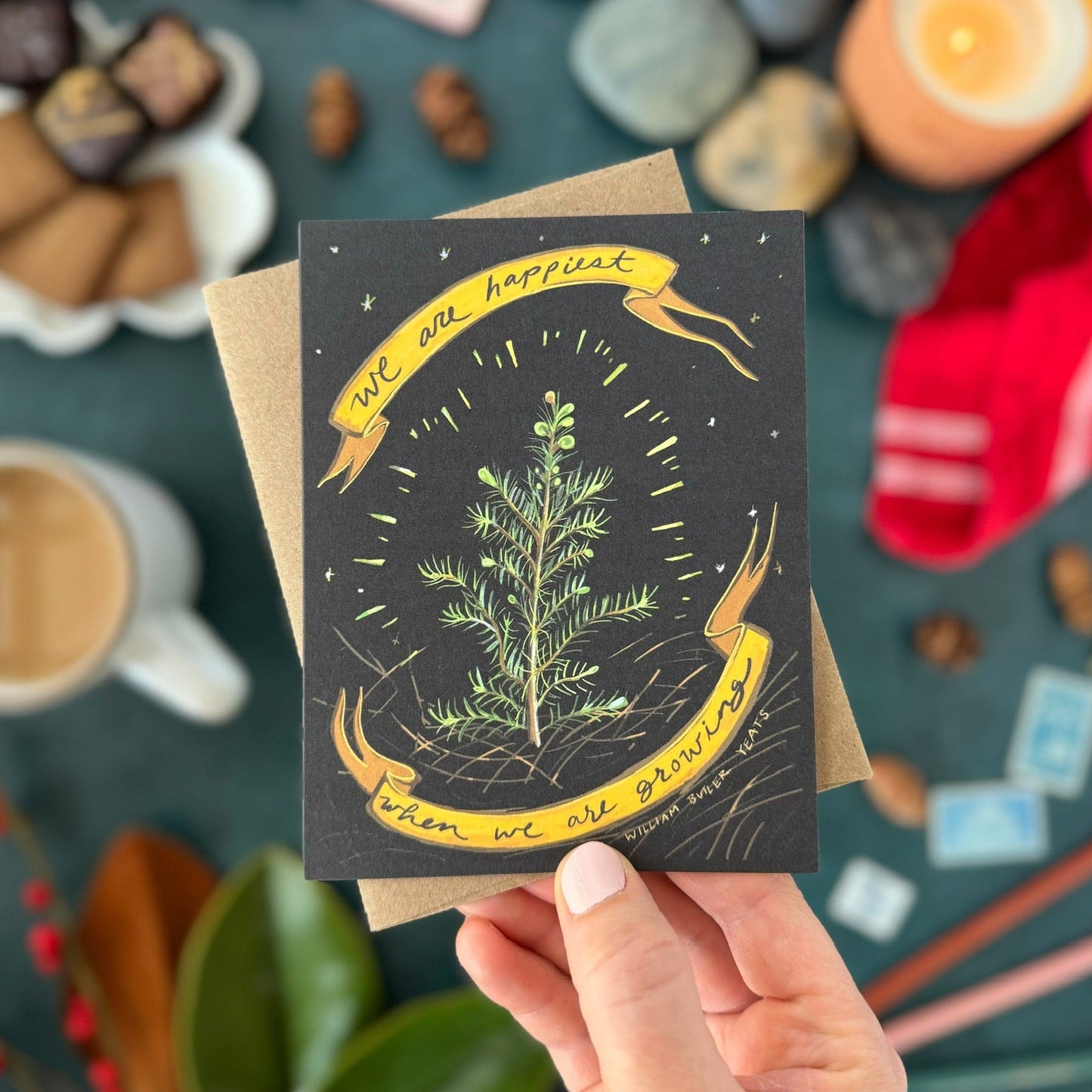 Hand holding a greeting card that says, "We are happiest when we are growing," with a plant illustration and text, surrounded by various items on a dark surface.