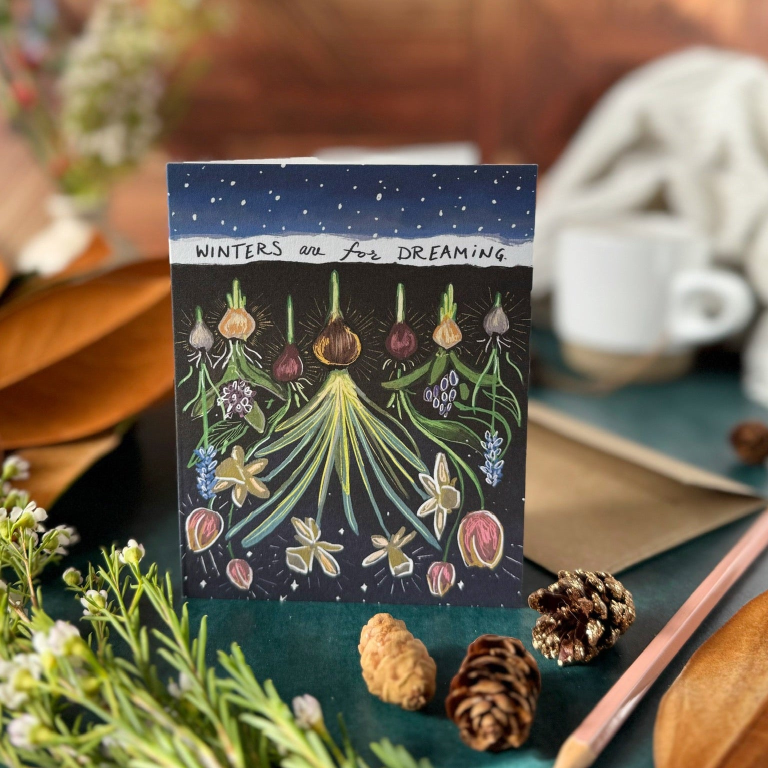 Greeting card with nature-themed design  that says "Winters are for dreaming," on a wooden surface with plants and a cup.
