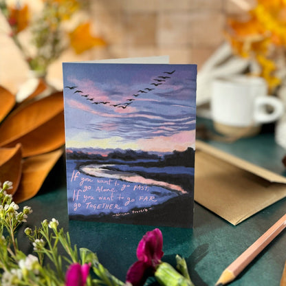 Greeting card with a scenic design of birds flying over water, placed on a table with flowers and a pencil.