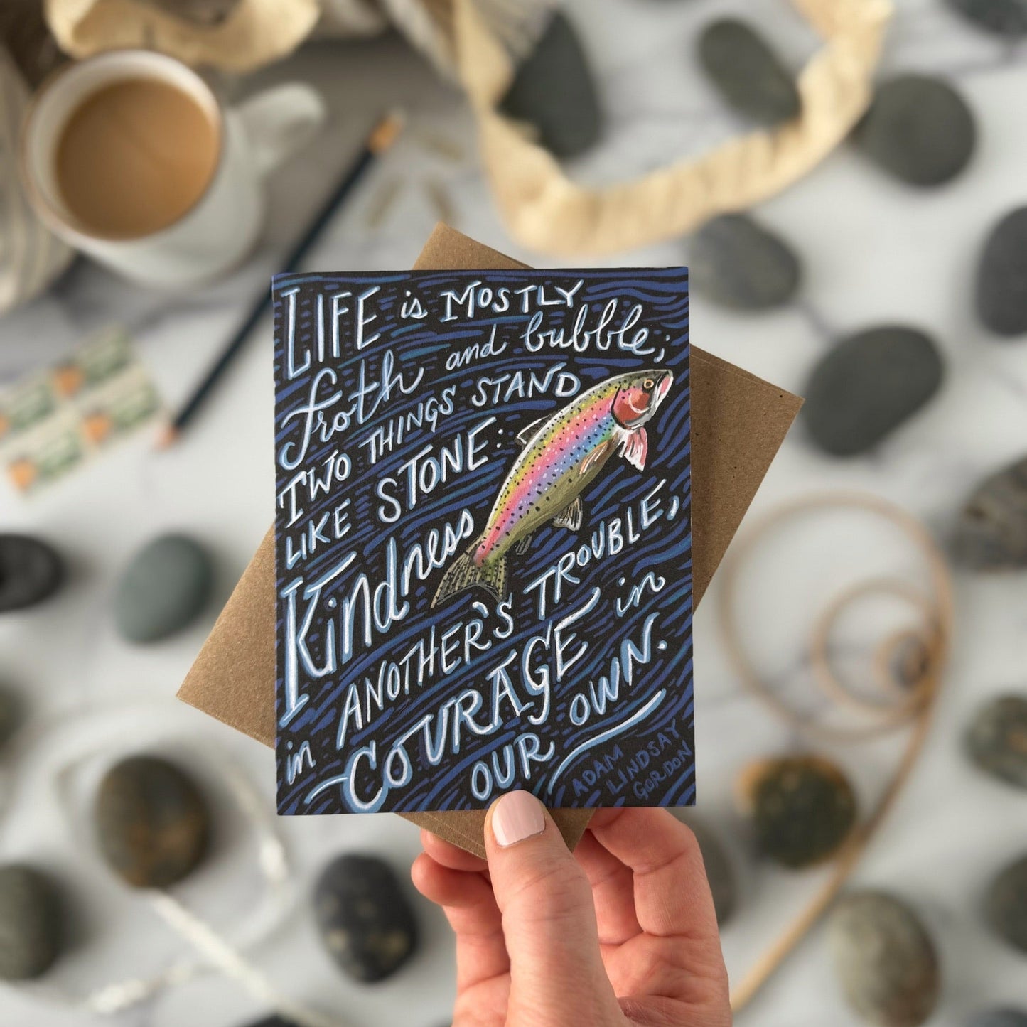Hand holding a card with a rainbow fish illustration and inspirational quote on a pebble background.