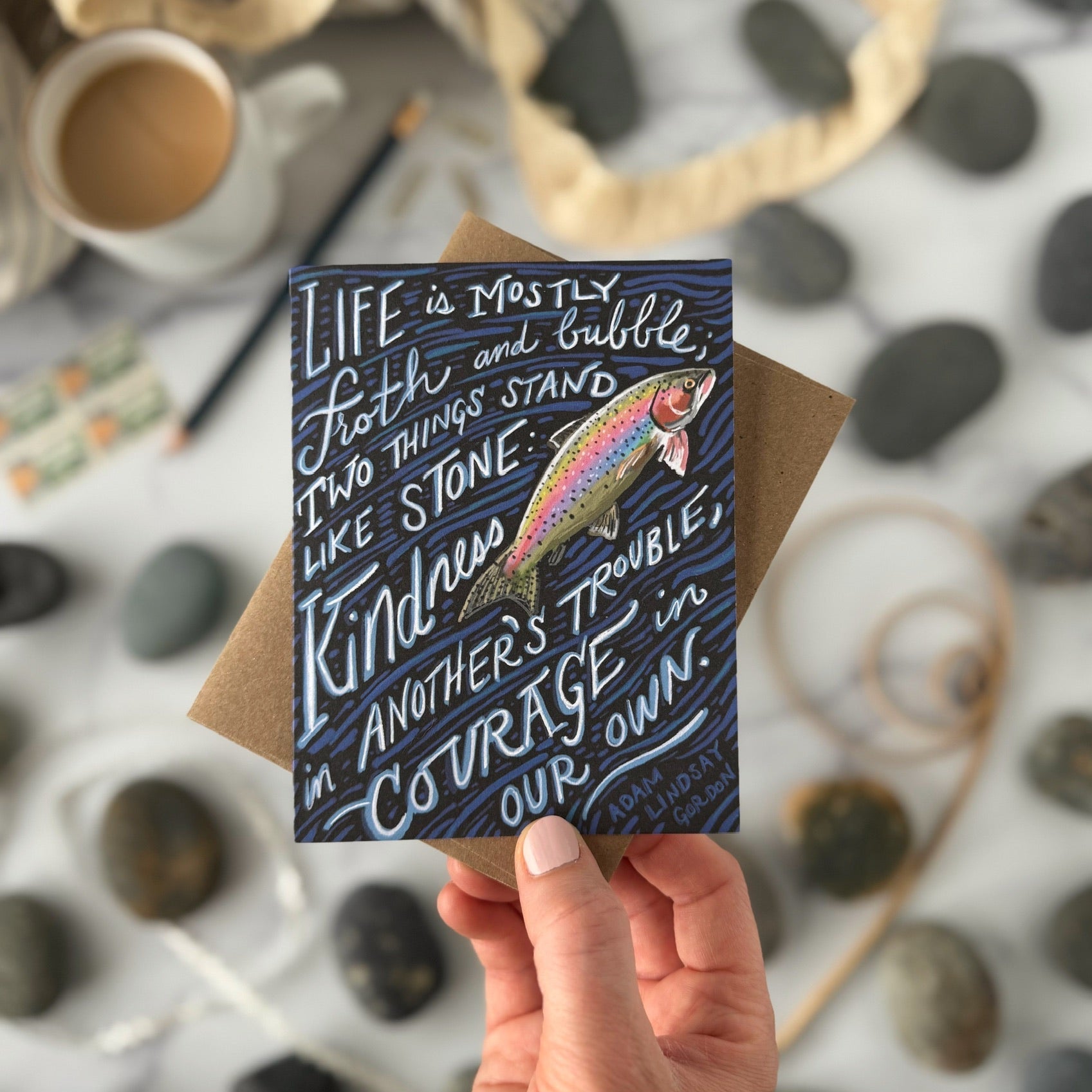 Hand holding a card with a rainbow fish illustration and inspirational quote on a pebble background.