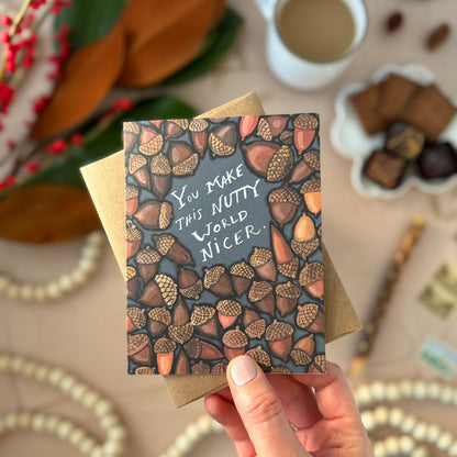 Hand holding a card with acorn design and text that says "You make this nutty world nicer," with a blurred background of coffee and nuts.