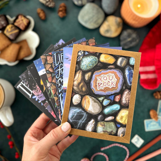 Hand holding a set of 8 greeting cards with the first card being a pebble design and 'You Rock' text, surrounded by snacks and decor on a dark surface.