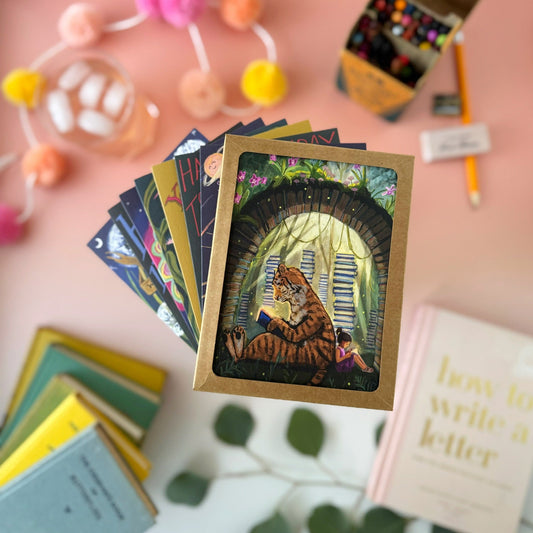 A fanned stack of illustrated art cards and one postcards on a pink tabletop, featuring a central card with a tiger and little girl reading under a book archway Surrounding props include books, a pencil, pom-poms, and a small box of colorful items.