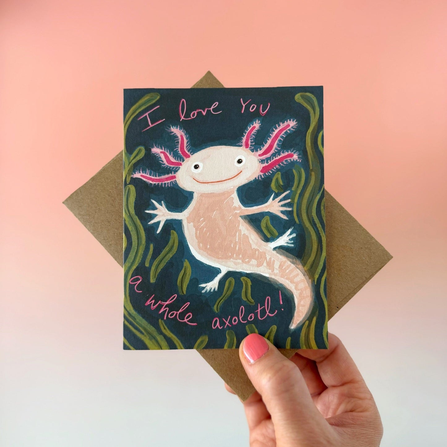 A hand holds a square greeting card featuring a whimsical axolotl illustration with the text 'I love you a whole axolotl!' on a dark background, with a brown envelope behind it against a soft pink-to-white gradient backdrop.