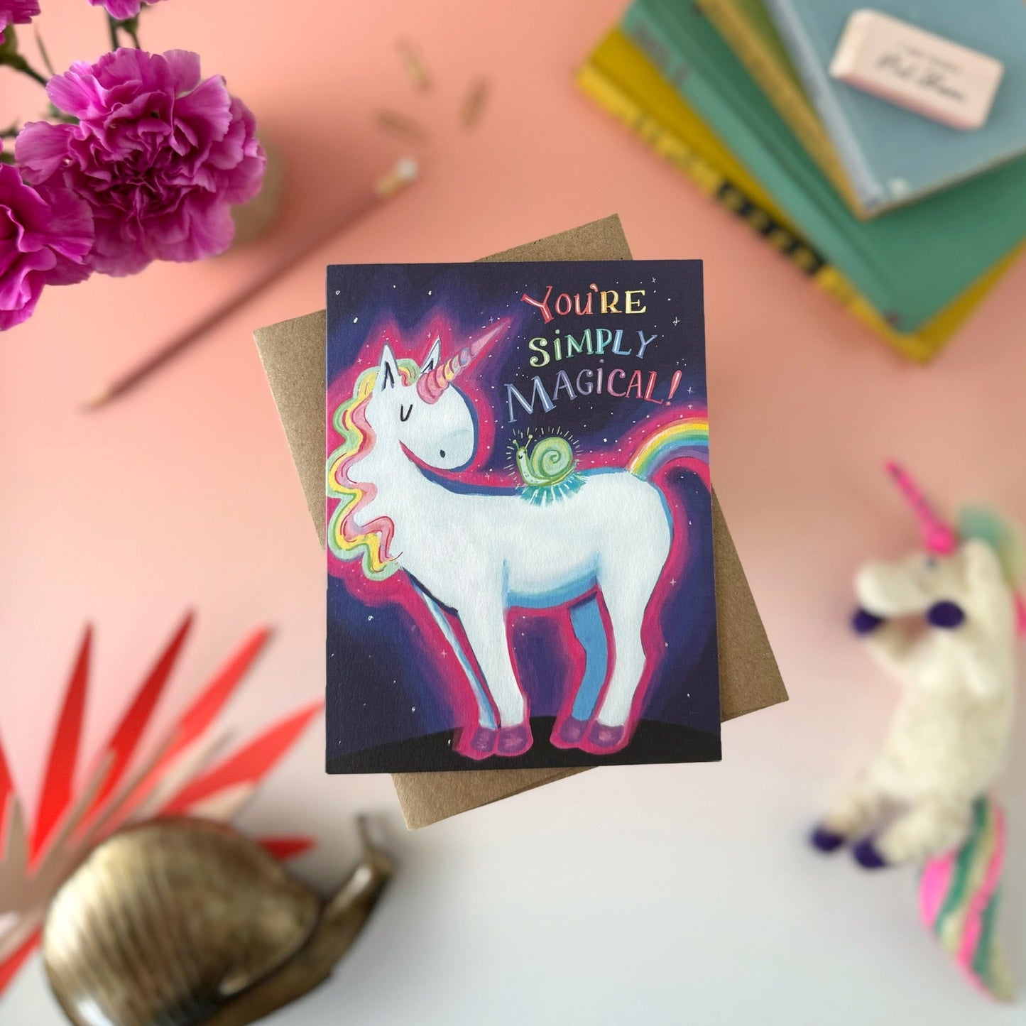 A square greeting card featuring a colorful unicorn illustration with the text 'You're Simply Magical!' on a dark starry background, styled on a kraft envelope with flowers, books, and a plush toy around it.