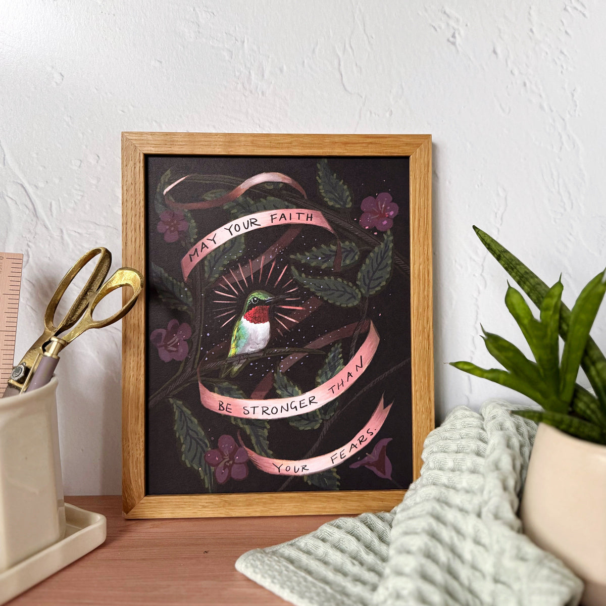 A framed art print featuring a hummingbird, pink banners with motivational text that says "May you faith be stronger than your fears", and green leaves on a dark background, styled on a wooden desk with a plant, scissors, and a textured towel.