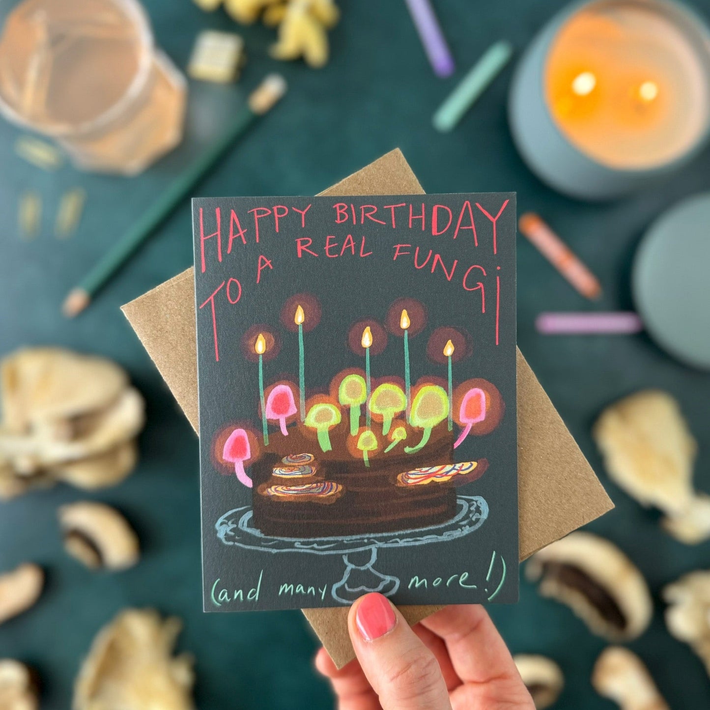 A hand holds a dark gray birthday card that reads 'HAPPY BIRTHDAY TO A REAL FUNGI' above an illustration of a mushroom cake with glowing candles, layered over a kraft envelope. The background is a styled flat-lay with mushrooms, colored pencils, and a lit candle on a dark teal surface.