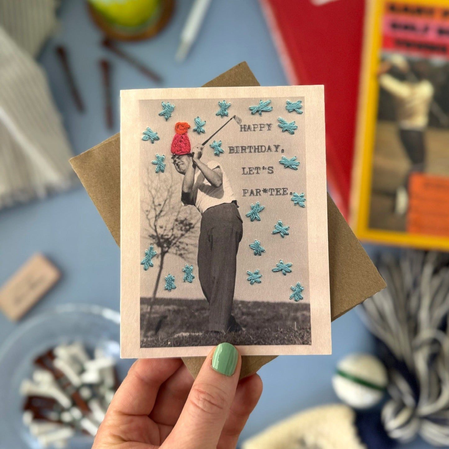 Hand holding a greeting card with a vintage-style photo of a person in a golf outfit, surrounded by books and stationery items.
