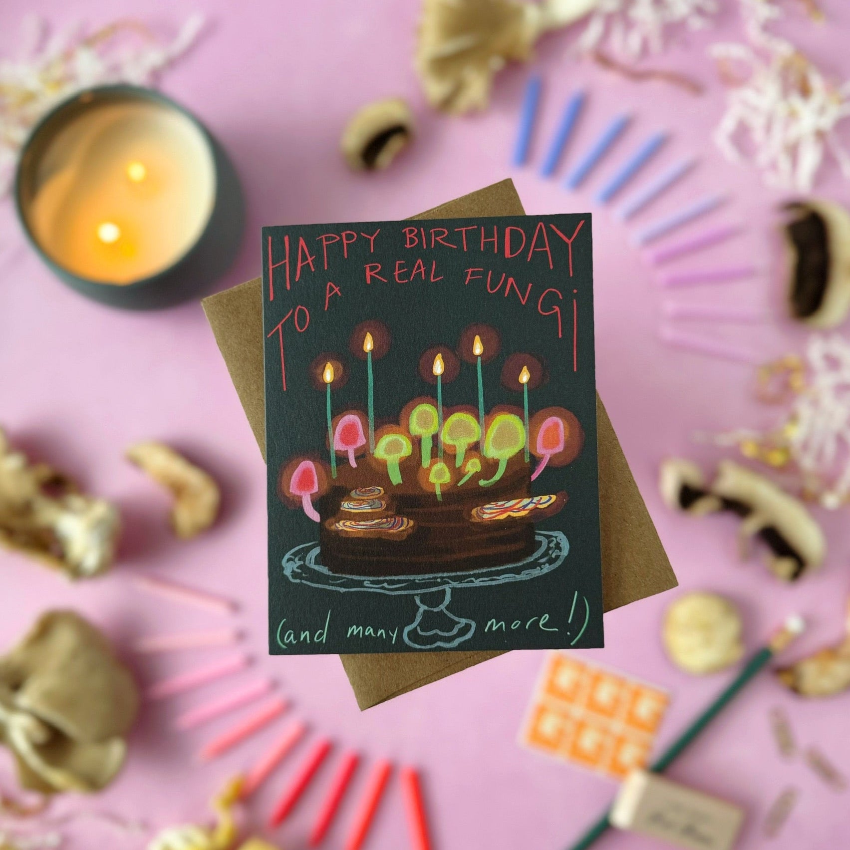 A birthday card with a dark background and colorful illustrated candles and cake with glowing mushrooms and turkey tail mushooms sprouting out of the cake. The text says "Happy Birthday to a real fungi (and many more)". The card sits on a kraft envelope, styled in a flat lay on a pink surface with dried mushrooms, candles, stamps, and small decorative objects around it.