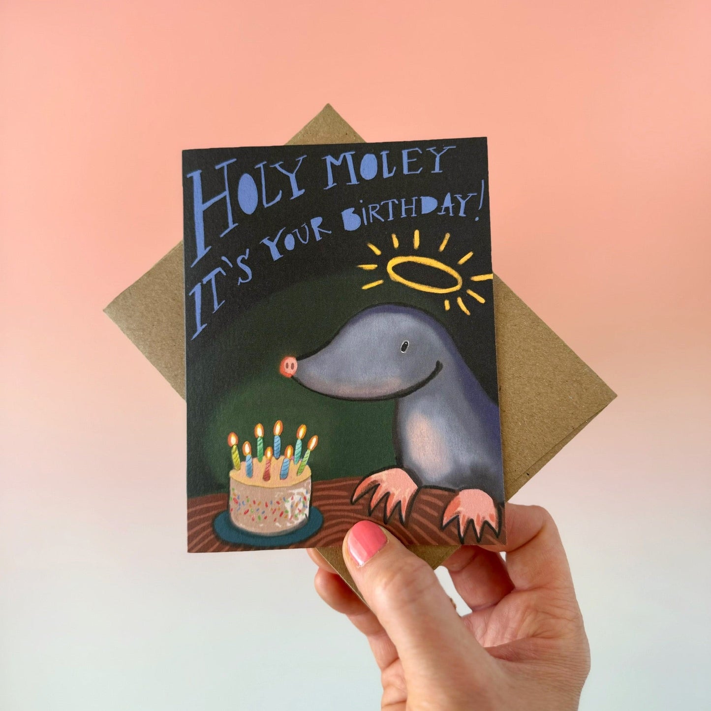 A hand holds a birthday card featuring a playful gray mole with a halo and a birthday cake, with the text 'HOLY MOLEY IT'S YOUR BIRTHDAY!'. The card is shown with a brown envelope against a soft pink-to-blue gradient background.