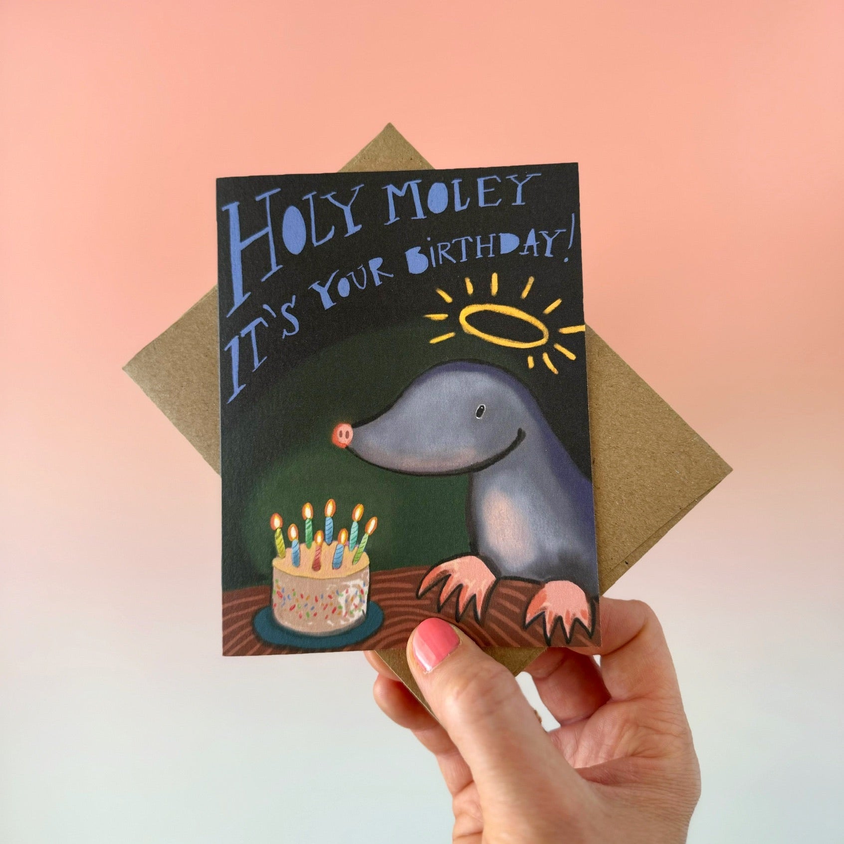 A hand holds a birthday card featuring a playful gray mole with a halo and a birthday cake, with the text 'HOLY MOLEY IT'S YOUR BIRTHDAY!'. The card is shown with a brown envelope against a soft pink-to-blue gradient background.