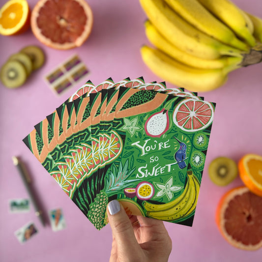 A hand holds a fan of illustrated fruit-themed postcards that say "You're So Sweet" on a pink surface, surrounded by real fruit props including bananas, grapefruit halves, kiwi slices, and a pen.