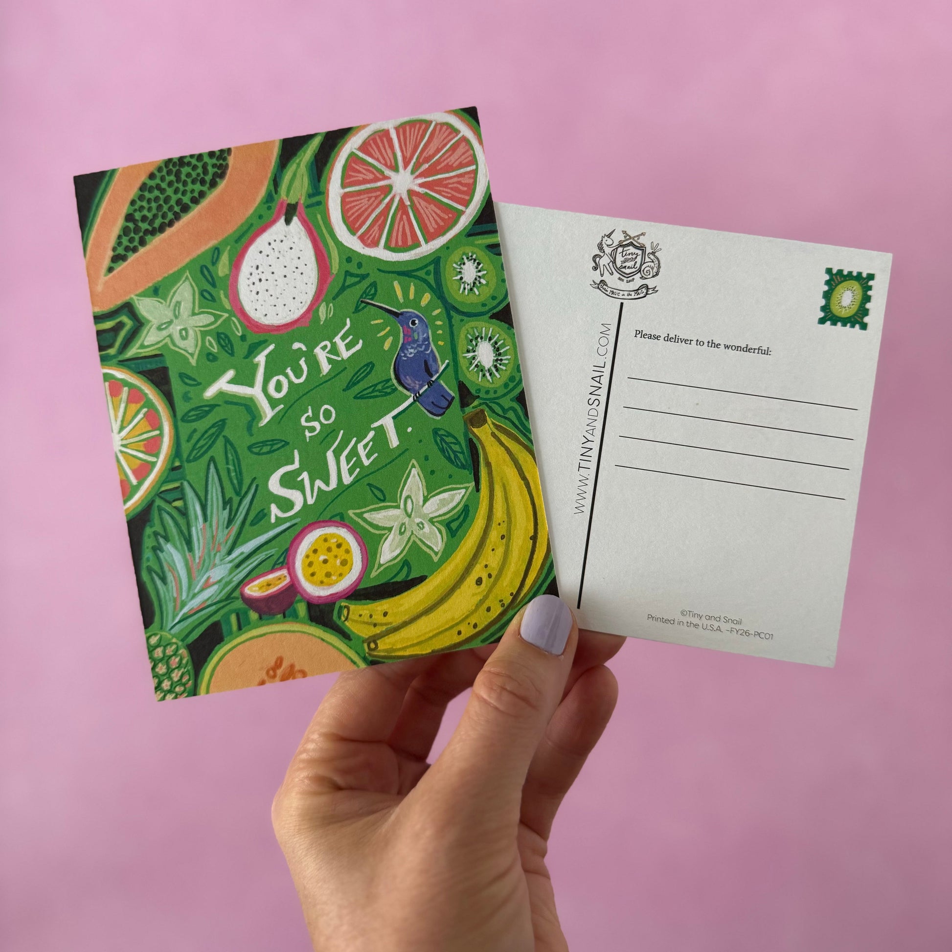 A hand holds a colorful illustrated postcard set against a solid pink background. The front of the postcard features a vibrant tropical fruit design with the text 'You're so Sweet' and a small hummingbird, while the back shows a clean white writing area with a postage stamp illustration.