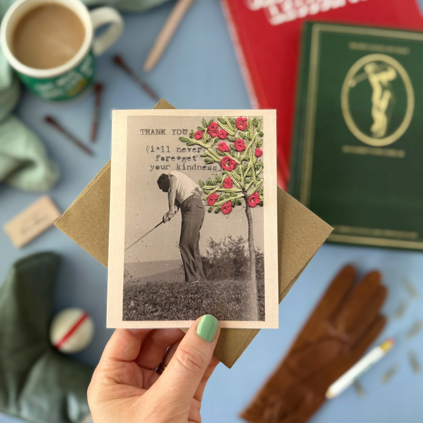 Hand holding a vintage-style card with a golfing scene and tree, surrounded by books and stationery on a blue surface.