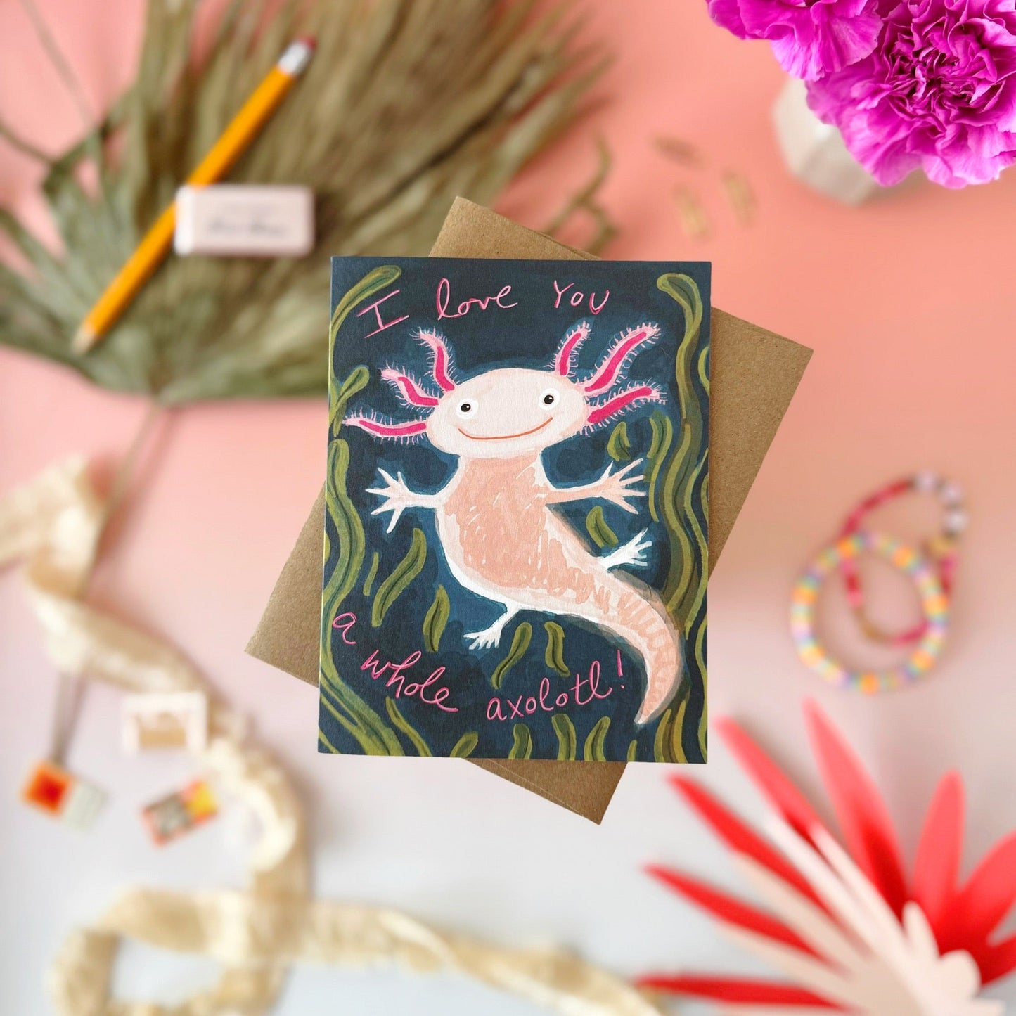 A greeting card featuring a cute hand-drawn axolotl on a dark blue-green background with pink and green details, placed on top of a brown kraft envelope. The card is photographed in a styled flat-lay arrangement on a pink surface with decorative props including flowers, a pencil, ribbon, and small accessories.