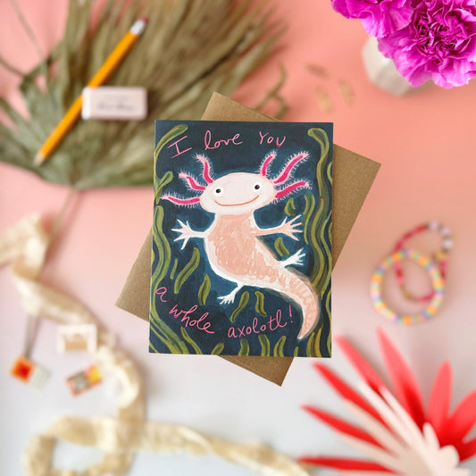 A greeting card featuring a cute hand-drawn axolotl on a dark blue-green background with pink and green details, placed on top of a brown kraft envelope. The card is photographed in a styled flat-lay arrangement on a pink surface with decorative props including flowers, a pencil, ribbon, and small accessories.
