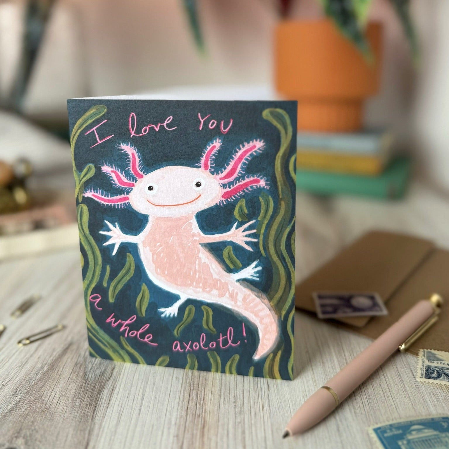 A greeting card featuring a hand-drawn pink axolotl on a dark blue-green background, placed on a light wood tabletop with a pen, stamps, and a potted plant in a blurred room behind it.