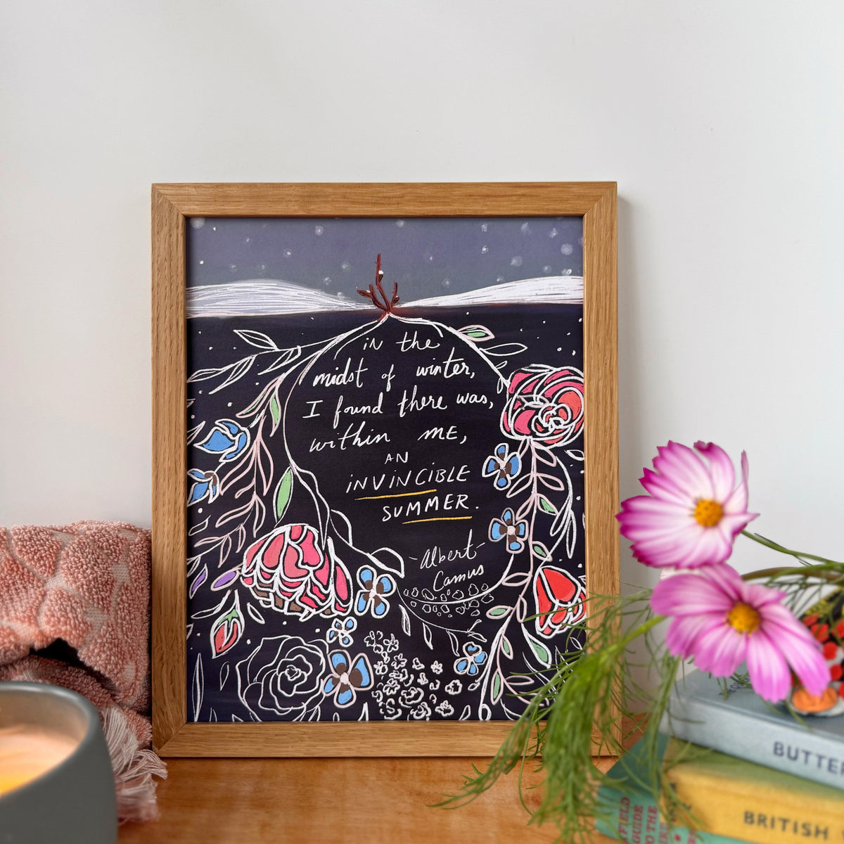 A framed art print featuring a handwritten quote: "In the midst of winter, I found there was, within me, an invincible summer. Albert Camus" Accompanied by colorful floral illustrations, styled on a wooden table with a pink blanket, a candle, and pink flowers.