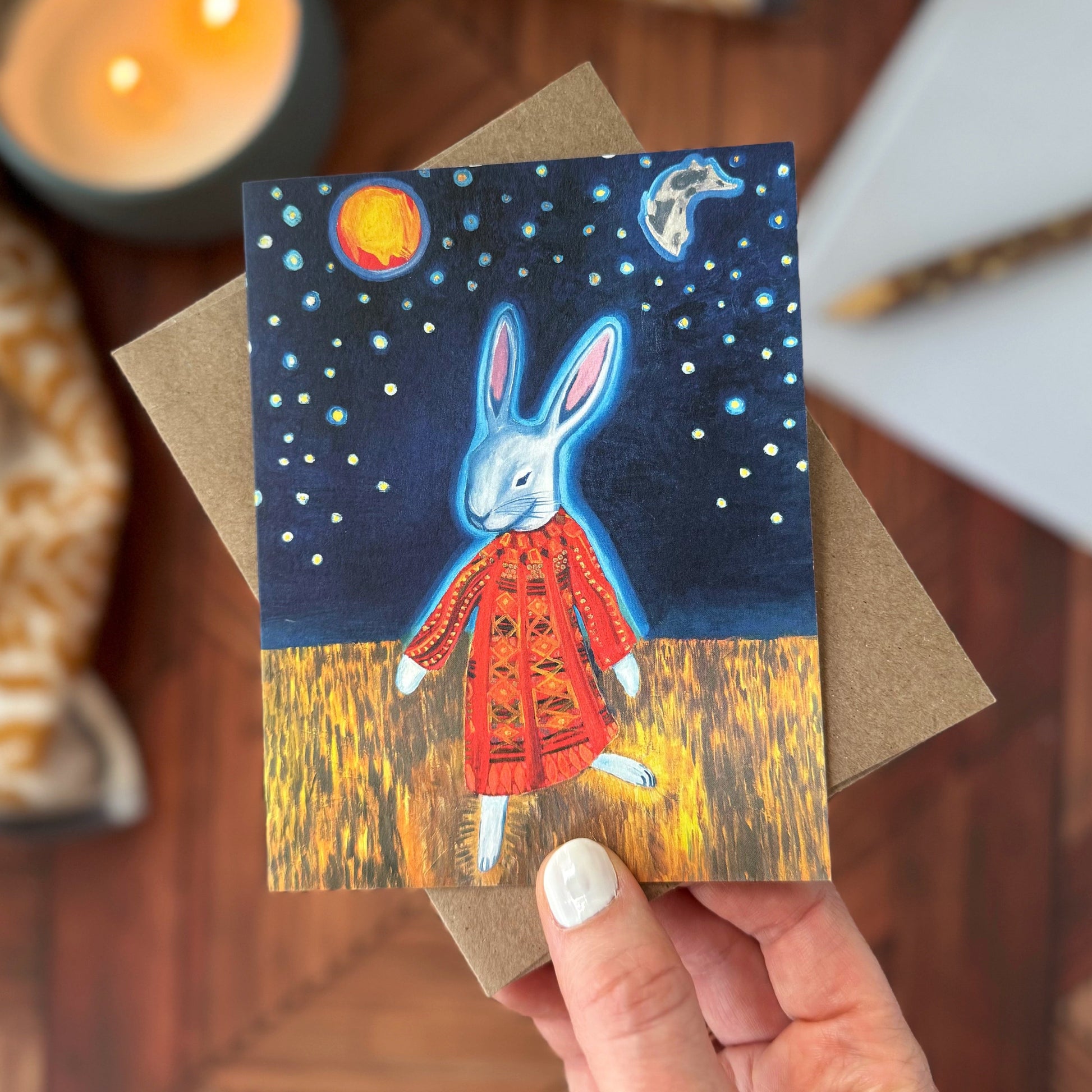A hand holds a greeting card featuring an illustrated white rabbit in a quilted red coat under a starry night sky, with a brown envelope behind it and a warm wooden tabletop around it.