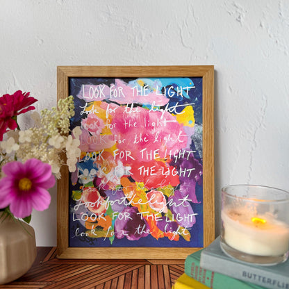 A framed art print featuring colorful watercolor splotches and white handwritten text reading 'LOOK FOR THE LIGHT' styled on a wooden table with a candle, books, and flowers against a textured white wall.