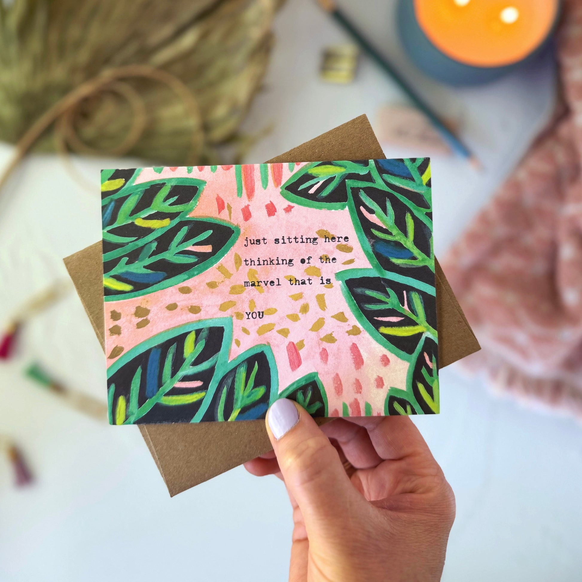 Hand holding a card with leaf design and text, surrounded by decorative elements on a light surface.