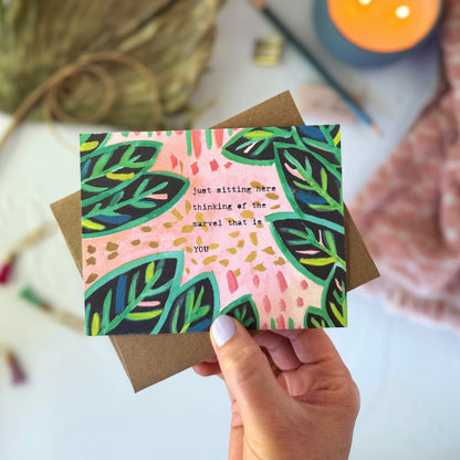 Hand holding a card with leaf design and text, surrounded by decorative elements on a light surface.