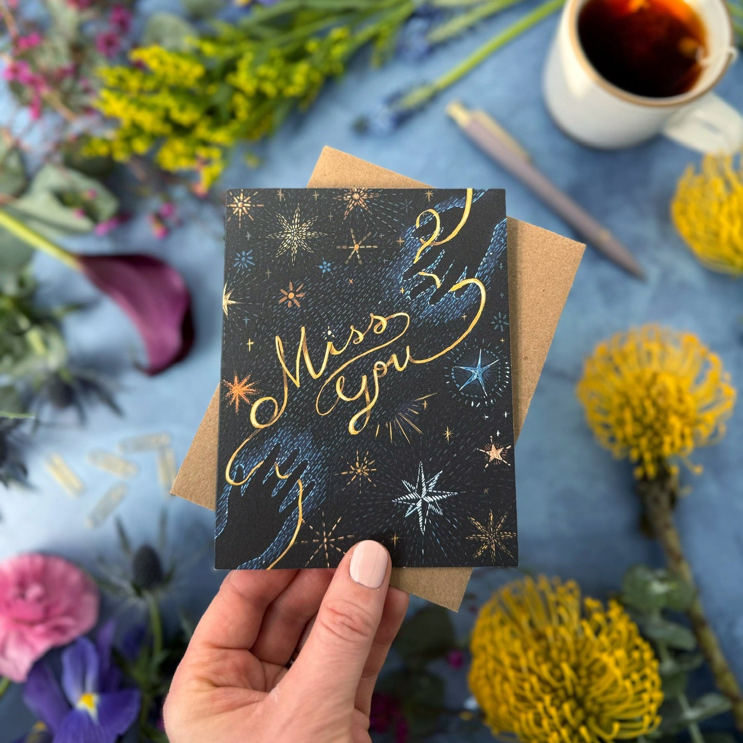 Hand holding a card with 'Miss You' text amidst flowers and a cup of tea.