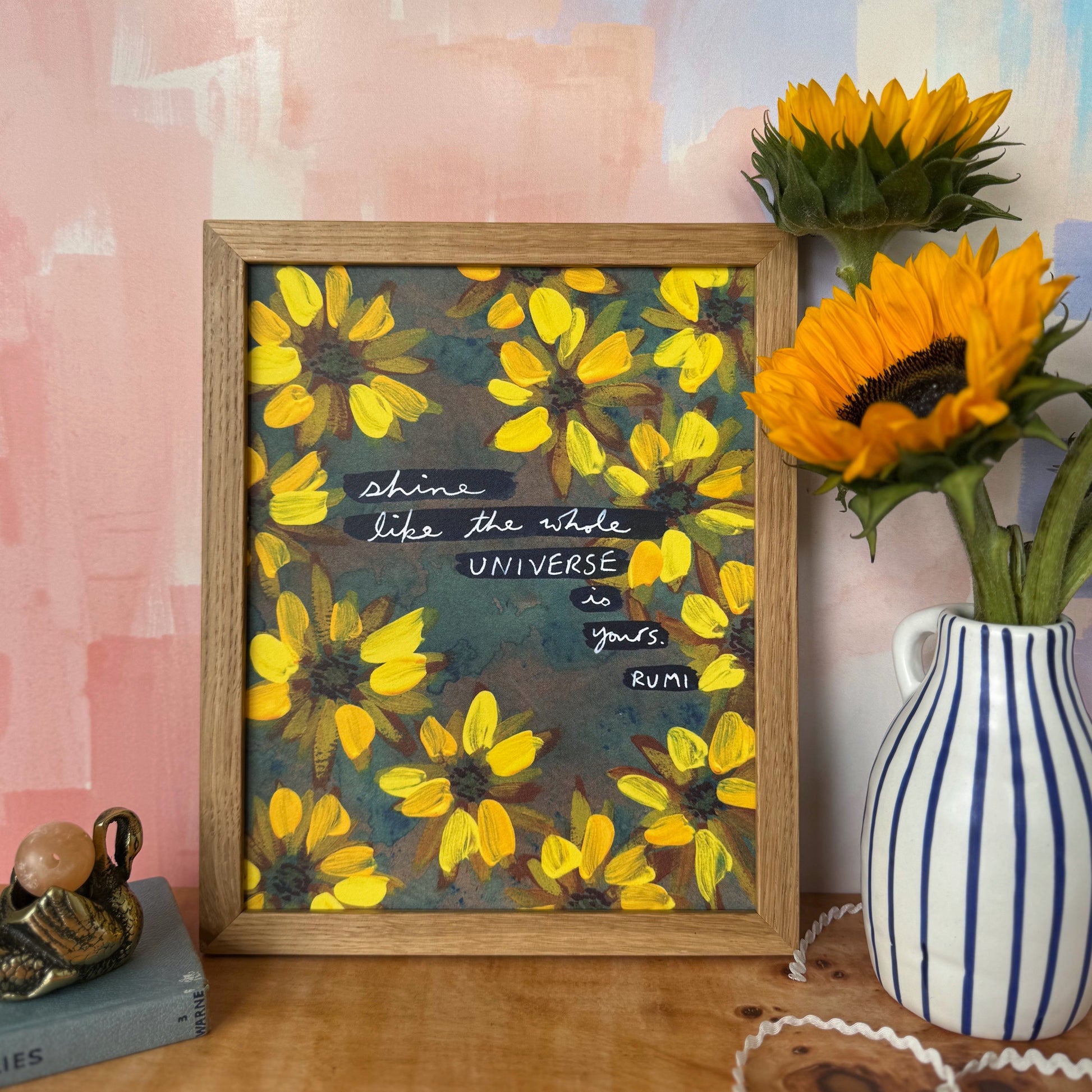 A framed art print featuring a yellow flower pattern and a quote by Rumi, styled on a wooden table next to sunflowers in a blue-and-white striped vase against a pink and blue painted wall.