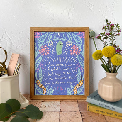 A framed art print featuring a whimsical purple and blue illustration with a green chrysalis, caterpillars,  flowers, and handwritten text, styled on a terracotta tile surface next to a vase of yellow flowers and desk accessories.