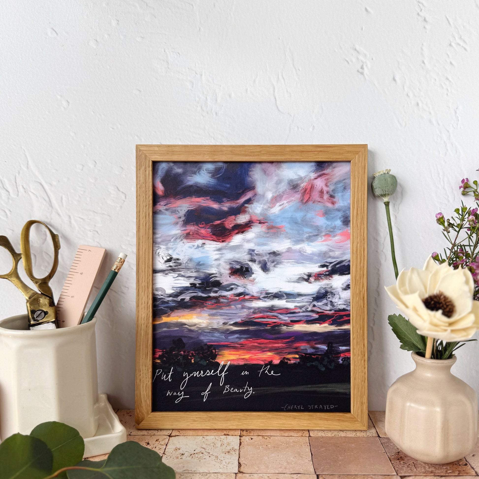 A framed art print featuring a dramatic sunset sky with pink, red, blue, and white clouds over a dark landscape, styled on a terracotta tile surface against a textured white wall with decorative props.