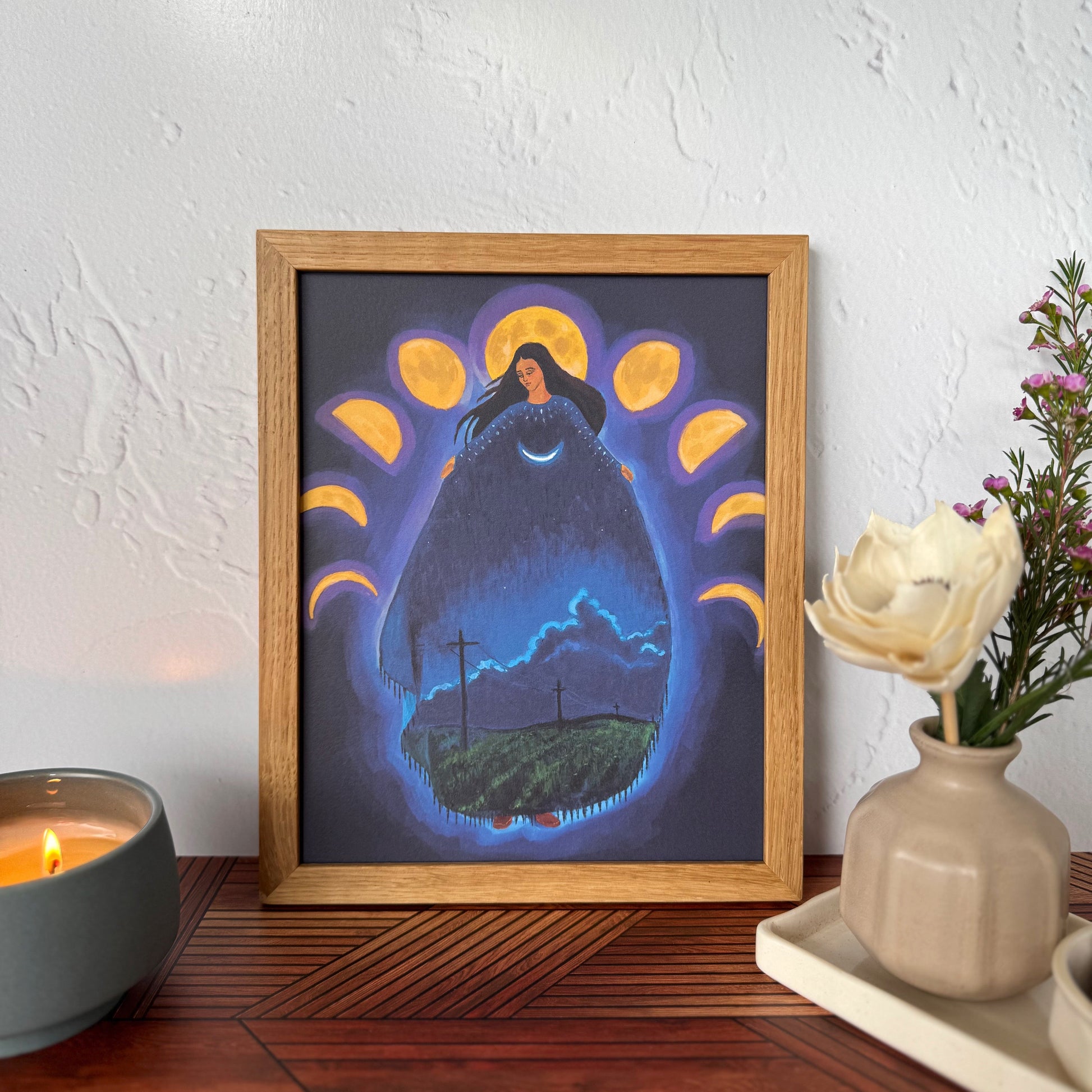 A framed art print featuring a surreal night scene with a glowing form of a woman surrounded by moon phases,  displayed on a wooden table against a textured white wall with a candle and flowers nearby.