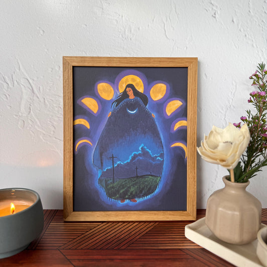 A framed art print featuring a surreal night scene with a glowing form of a woman surrounded by moon phases,  displayed on a wooden table against a textured white wall with a candle and flowers nearby.