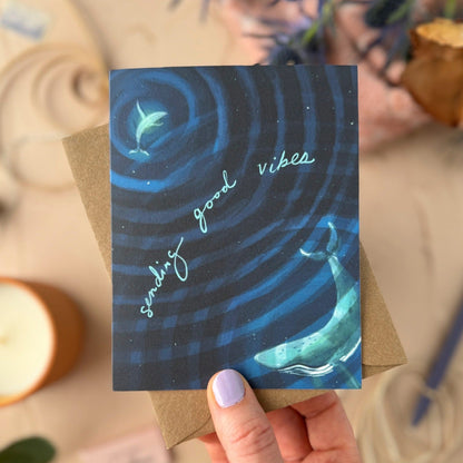 Hand holding a card with whale design and 'sending good vibes' text, surrounded by candles and plants.
