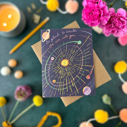 A greeting card with a solar system illustration lies on top of a brown kraft envelope, styled in a flat lay with flowers, pom-poms, a candle, and a pencil on a dark teal surface.
