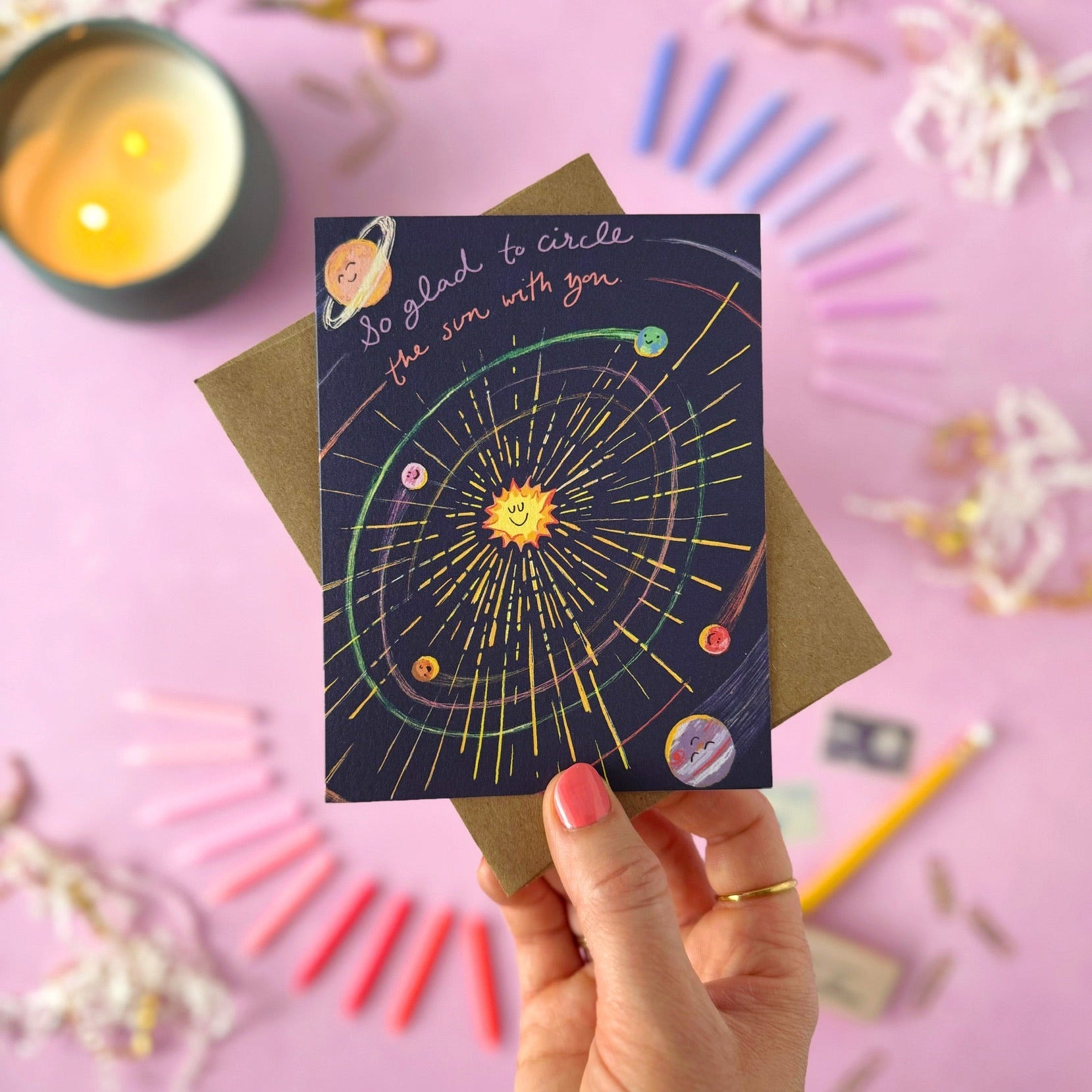 A hand holds a solar system themed greeting card that says "So glad to circle the sun with you"  with a brown envelope, set against a pink background decorated with stationery and a candle.