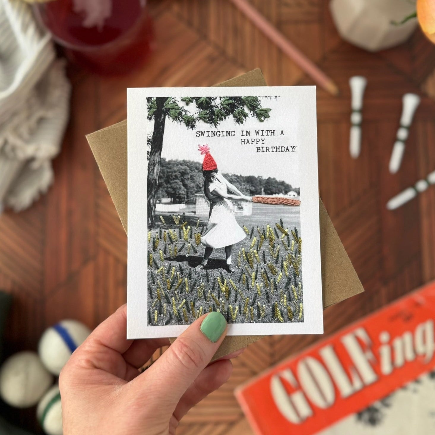 Hand holding a greeting card with a vintage-style photo of a woman golfing in a field, surrounded by a casual setting with a drink and flowers.