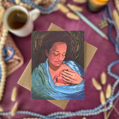 A greeting card featuring a painted portrait of a woman in a blue robe holding a baby, placed on a kraft envelope over a purple surface with a cup, candle, pencil, and decorative beads around it.