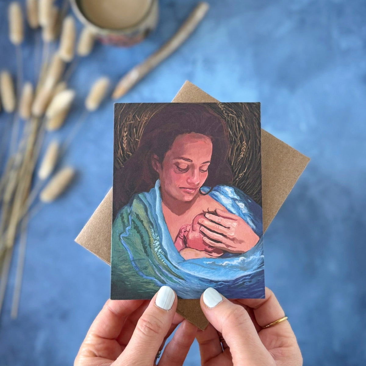 A hand holds a greeting card showing a painted scene of a woman cradling a baby, with a brown envelope behind it on a textured blue surface. A mug, dried reeds, and a pencil are softly blurred in the background.